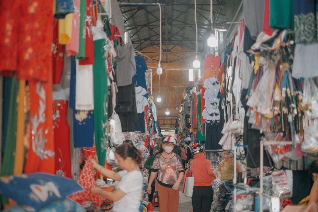 A bustling indoor market with diverse clothing items on display and shoppers exploring.